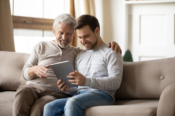 Smiling mature father and grownup son relax on sofa in living room have fun using modern tablet...