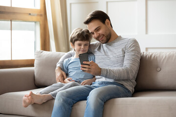 Smiling young father and little son sit relax on couch in living room have fun using cellphone together. Happy dad and small boy child rest on sofa at home watch funny video on smartphone gadget.
