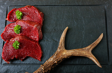 Raw steak meat from roe deer on the bridlic  chopping board. Roe deer antler as a decoration. Copy...