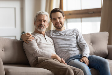 Family portrait of happy millennial Caucasian man with elderly 70s father sit relax on couch at home. Smiling mature dad with adult grownup son rest on sofa, enjoy weekend together. Bonding concept.