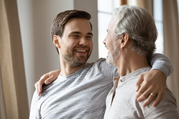 Overjoyed millennial Caucasian man and elderly dad hug enjoy family weekend at home together. Smiling adult grownup son embrace mature father, show love and care. Gratitude, bonding concept.
