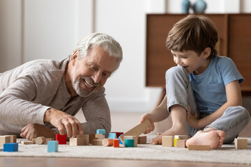 Obraz na płótnie Canvas Loving elderly Caucasian grandfather have fun playing with little 6s boy child building with blocks. Happy caring mature grandparent engaged in funny game activity with small grandson at home.