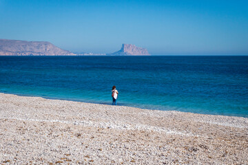 Girl alone on the stone beach of Albir with view over ocean to rock of Calpe, Albir, Spain