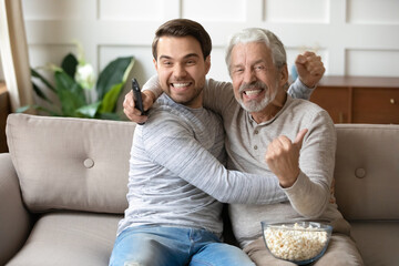 Overjoyed young Caucasian man with senior father relax at home on couch cheer support watching...
