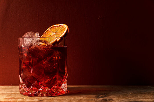 Glass with ice cocktail type americano or negroni with whole orange slice on red wall background and ray of light
