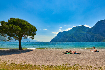 Riva del Garda,Lago di Garda ,Italy  - 12 June 2020: People who bathe and sit on the beach to tan at Lake Garda, beautiful Lake Garda surrounded by mountains in the summer time