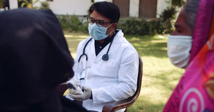 Slow-motion handheld shot of male doctor with face mask giving advice suggestion for precautions  and safety outdoors to females, one in red saree and another in black hijab during global pandemic   
