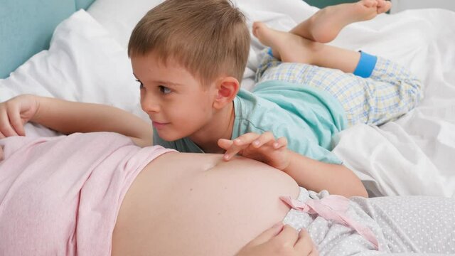Happy smiling toddler boy in pajamas stroking big belly of pregnant mother and talking to her. Concept of loving children and family happiness expecting baby