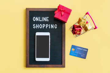 Miniature of gift boxes in trolley, mobile phone, word on black frame and credit card on yellow background. Online shopping concept. Top view, flat lay and copy space