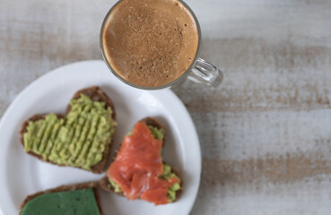 Sandwich with avocado paste, green cheese with basilic and salmon in the shape of heart, cup of coffee