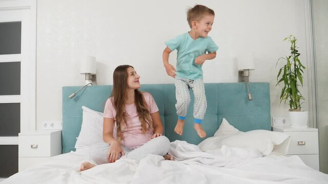 Beautiful young woman in pajamas looking on her little toddler son jumping on her bed at morning. Concept of cheerful children and family happiness.