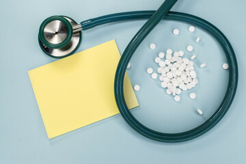 A stethoscope with white pills on a blue background next to a yellow blank sheet of paper. The concept of medical pharmacy. Banner for the consumption of painkillers or prescription drugs