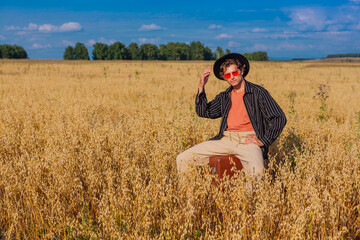 Tall handsome man with black hat and pink sunglasses sitting on a brown vintage leather suitcase at golden oat field.