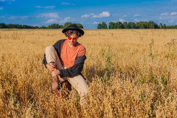 Naklejka na ściany i meble Tall handsome man with black hat and pink sunglasses sitting on a brown vintage leather suitcase at golden oat field.