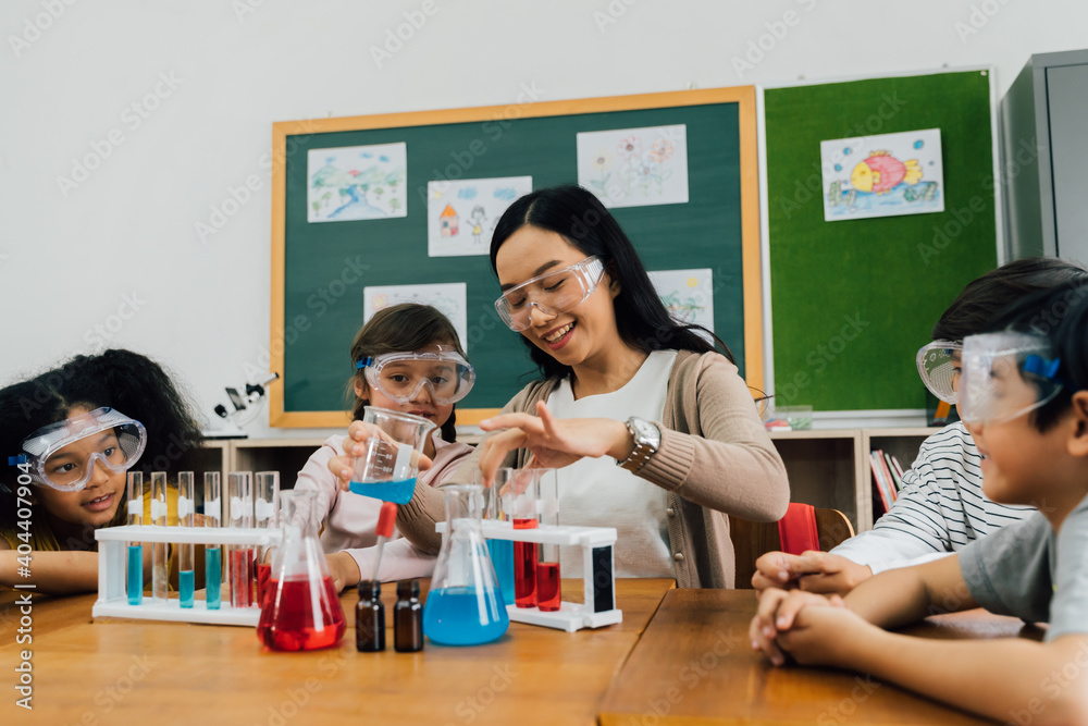 Wall mural Young woman mixing liquids in test tube with multiethnic children watching, learning about science, chemistry. Asian school teacher and students in science class