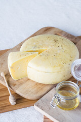Homemade cheese wheel with cutted piece on cutting boards, honey in glass jar and honey spoon on textile background, selective focus