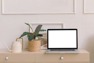 Obraz na płótnie Canvas Laptop computer with blank screen, home plant, watering can, unrecognizable picture in frame on dresser. Home interior details in pastel beige colors.