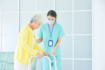 Nurse wearing face mask helping senior woman patient to walk by walker at nursing home.