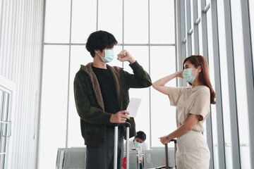 Fototapeta na wymiar Asian young tourists wearing face mask greeting each other with their elbows at airport terminal, people sitting distance away from each other to prevent covid19 infection, red cross shows on empty ch