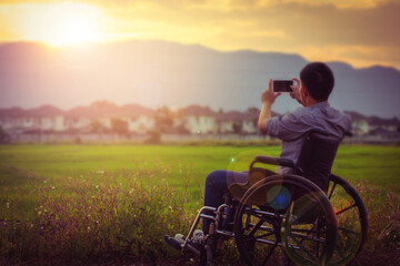Fototapeta na wymiar Close up of a paralyzed man sitting on wheelchair took a photo of the sunset by smartphone at rice green field with the blurred village and mountain background in the evening 
