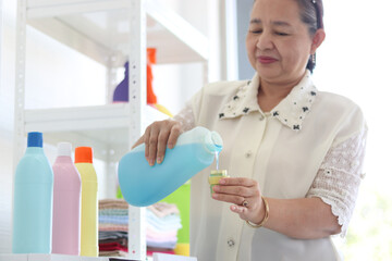Happy smiling Asian senior elderly woman housewife pouring fabric softener for doing laundry at laundry room with washing machine, grandma doing housework and cleaning clothes