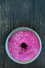 Vertical picture of bright pink donut on white round plate. Aged wooden background. Isolated object close up. Traditional American sweets for breakfast. Copy space. White sugar decoration on top. 