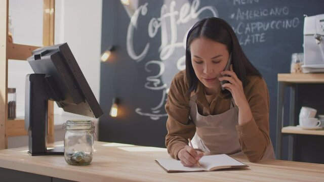 Panning medium close-up of young concentrated asian woman leaning over cashier desk talking on phone while doing paperwork owning small indoor coffeeshop