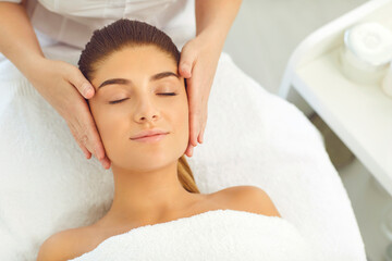 Fototapeta na wymiar Facial massage. Hands of cosmetologist massaging relaxed womans face in spa beauty salon