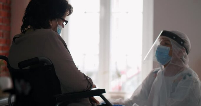 Medical support at the old woman's address. An old grandmother in a wheelchair is receiving mental therapy from a nurse. Full protective suit against covid-19