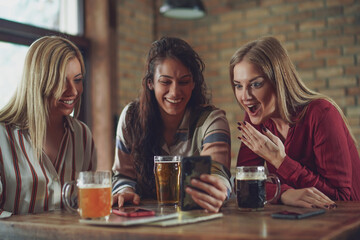 Three happy girlfriends are having fun time in pub. They are talking and drinking beer.