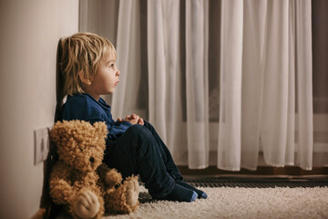 Sad little toddler child, blond boy, sitting in corner with teddy bear, punished