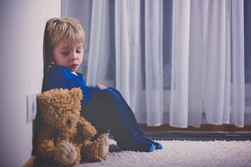 Sad little toddler child, blond boy, sitting in corner with teddy bear, punished