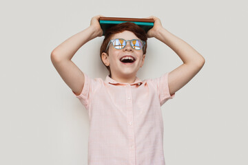 Happy ginger boy with glasses is holding some books on head and smile on a white studio wall