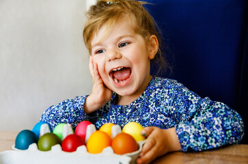 Fototapeta na wymiar Excited little toddler girl coloring eggs for Easter. Child looking surprised at colored egg hoarding and celebrating catholic and christian holiday with family. Cute kid helping to color, indoors.