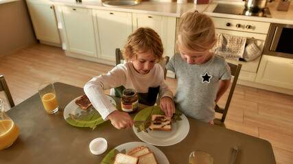 Loving brother teaching his little sister to spread chocolate nut butter on toasted bread while preparing breakfast in the kitchen