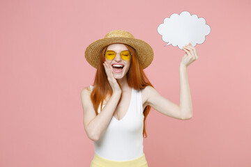 Fototapeta na wymiar Young laughing redhead woman 20s in straw hat glasses summer clothes hold empty blank say cloud, speech bubble for promotional content touch face isolated on pastel pink background studio portrait.