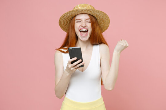 Young gladden redhead woman 20s in straw hat summer clothes hold in hand mobile cell phone doing winner gesture shout closed eyes clenching fists isolated on pastel pink background studio portrait