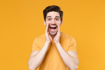 Young surprised shocked excited unshaved caucasian man 20s wearing casual basic blank print design t-shirt screaming shouting with hand on mouth isolated on yellow color background studio portrait