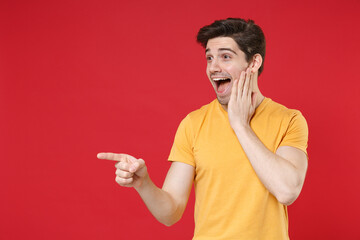 Young shocked unshaved man wearing casual yellow t-shirt pointing index finger aside on workspace copy space area put hand on face looking aside isolated on red color background studio portrait.