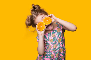 Cute girl wearing a dress is covering her eyes with sliced pieces of orange posing on a yellow wall