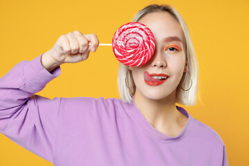 Young blonde funny caucasian woman 20s bob haircut bright makeup wearing casual basic purple shirt holding covering eye pink candy lollipop with tongue isolated on yellow background studio portrait.