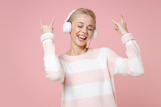 Young meloman woman dental braces short haircut in sweater listen music in headphones horns up gesture, depicting heavy metal rock sign, rock-n-roll isolated on pastel pink background studio portrait.