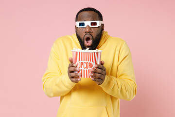 Shocked worried young african american man 20s in yellow streetwear hoodie 3d glasses watching movie film hold bucket of popcorn keeping mouth open isolated on pink color background studio portrait.