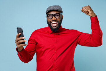 Joyful young bearded african american man 20s in casual red shirt eyeglasses cap doing winner gesture using mobile cell phone typing sms message isolated on pastel blue background studio portrait.