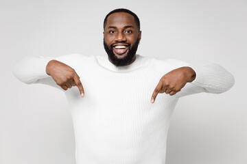 Funny cheerful smiling young african american man 20s wearing casual basic sweater standing pointing index fingers down on mock up copy space isolated on white color wall background studio portrait.