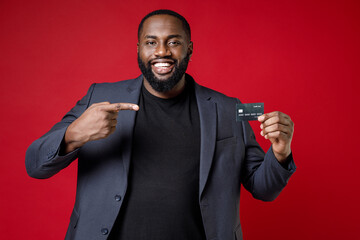 Smiling handsome young african american business man 20s wearing classic jacket suit standing pointing index finger on credit bank card looking camera isolated on red color background studio portrait.