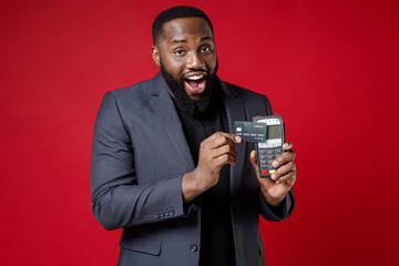 Shocked young african american business man 20s in classic jacket suit hold wireless modern bank payment terminal to process acquire credit card payments isolated on red background studio portrait.