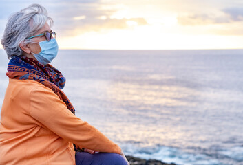 A senior grey-haired woman enjoying the outdoors and the horizon over water. Sitting on the cliff in front to the sea in a winter cloudy day, wearing surgical mask due to coronavirus