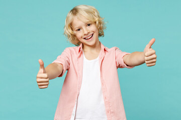 Cheerful little curly kid boy 10s in pastel pink shirt showing thumbs up like gesture looking...