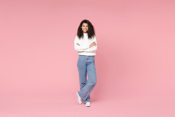 Full length young smiling african american woman 20s curly hair wear white casual knitted sweater jeans looking camera hold hands crossed folded isolated on pastel pink background studio portrait. - obrazy, fototapety, plakaty